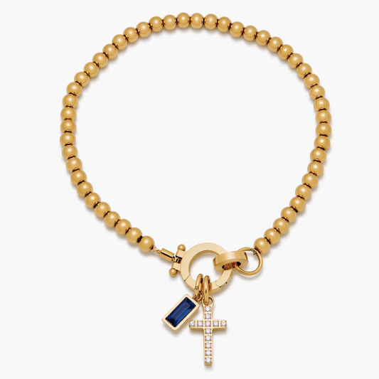 Pulsera de Veska con dije de cruz y piedra natal, hecha de metal y piedra colorida, con diseño elegante y simbólico.